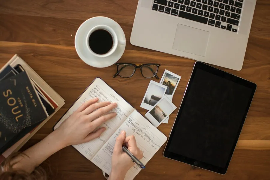 Persona escribiendo a mano en un diario junto a dispositivos electrónicos